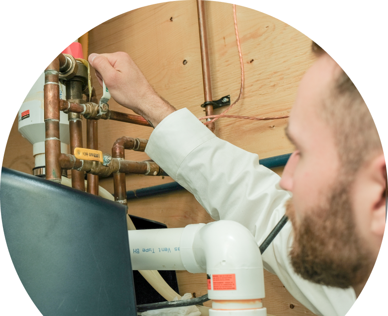 Shines water heater specialist tightens a valve on copper piping during a water heater installation, with exposed wood paneling and wiring in the background. Shines water heater specialist tightens a valve on copper piping during a water heater installation, with exposed wood paneling and wiring in the background.